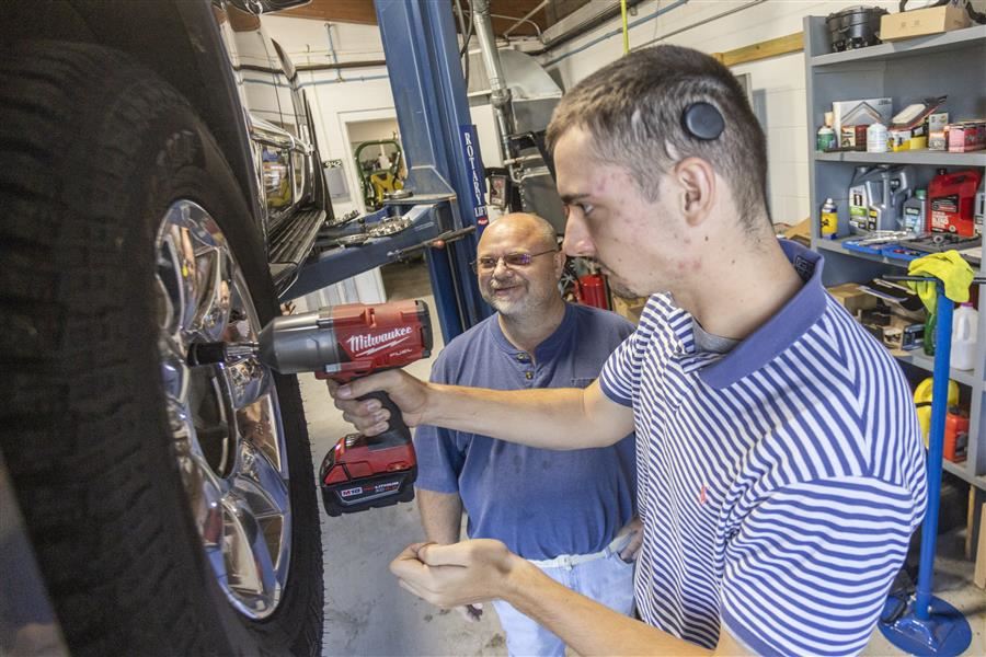 A student works to put a tire on a car. His instructor looks on proudly.